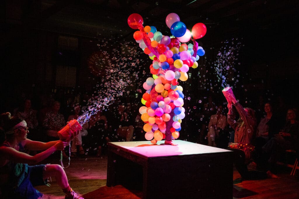 A performer on the 4x4 stage dressed in a full body costume made of balloons being shot at by bubbles